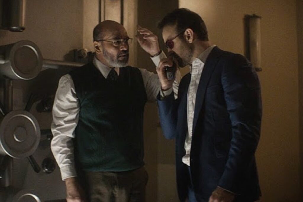 balding bearded man in glasses and a dress shirt and suit vest talking to a blind man with a cane