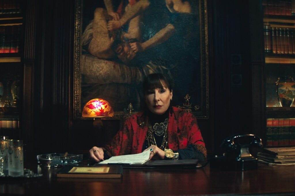 older dark haired woman in red sitting at a desk with a painting behind her