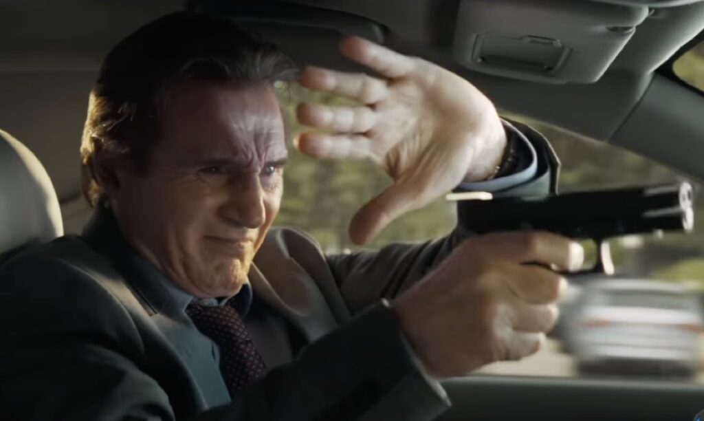 Liam Neeson trying to fire a gun through the front windshield of a car, one hand shielding his eyes.