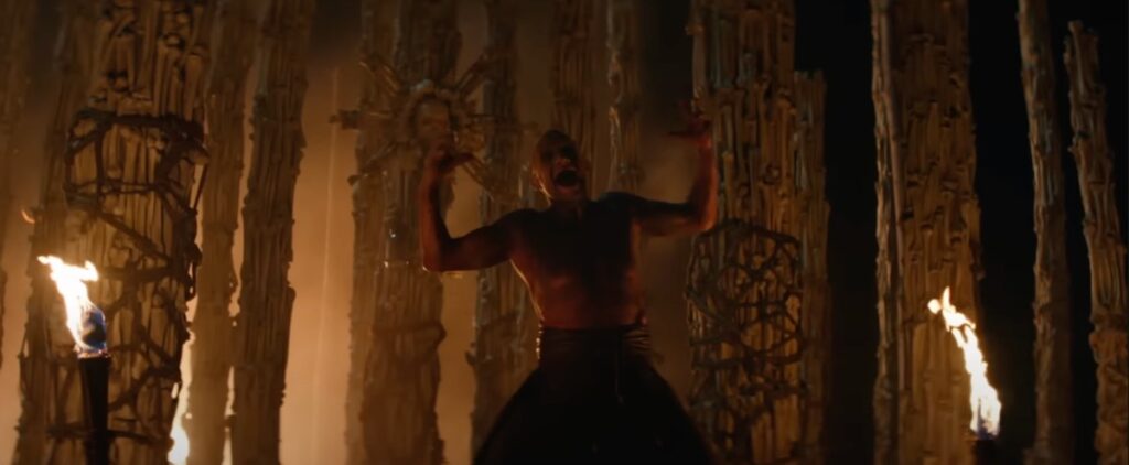 A bare-chested middle-aged man standing defiantly in the center of a bone temple.